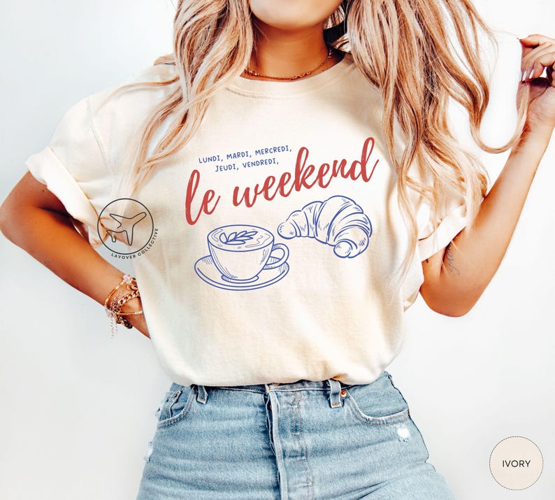May include: Ivory-colored t-shirt with the phrase "le weekend" in red script, accompanied by a blue line drawing of a coffee cup and croissant. The shirt also features the days of the week in French.