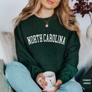 May include: A woman wearing a forest green sweatshirt with the text "NORTH CAROLINA" in white lettering. She is holding a white mug with floral designs.