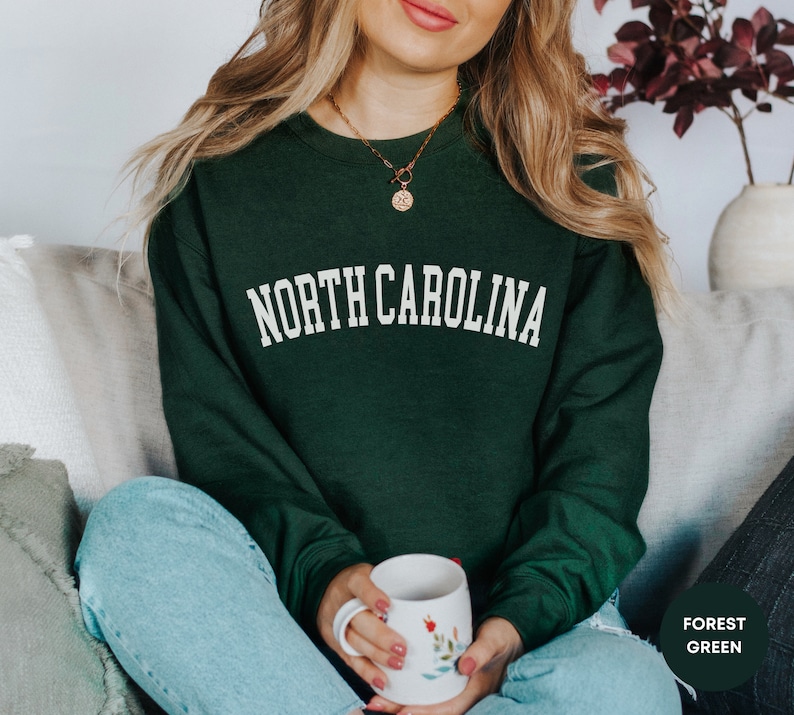 May include: A woman wearing a forest green sweatshirt with the text "NORTH CAROLINA" in white lettering. She is holding a white mug with floral designs.
