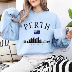 Puede incluir: Sudadera azul claro con la palabra "PERTH" en mayúsculas negras sobre una silueta del horizonte de Perth y la bandera australiana. La palabra "AUSTRALIA" está impresa debajo.