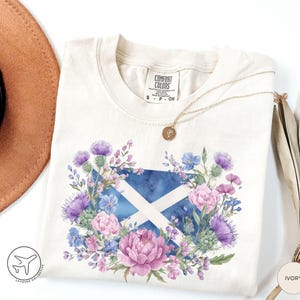 May include: Ivory-colored t-shirt featuring a watercolor design of the Scottish flag, surrounded by purple thistles, pink peonies, and blue flowers. A gold necklace and a brown hat are visible. The shirt has a Comfort Colors label.