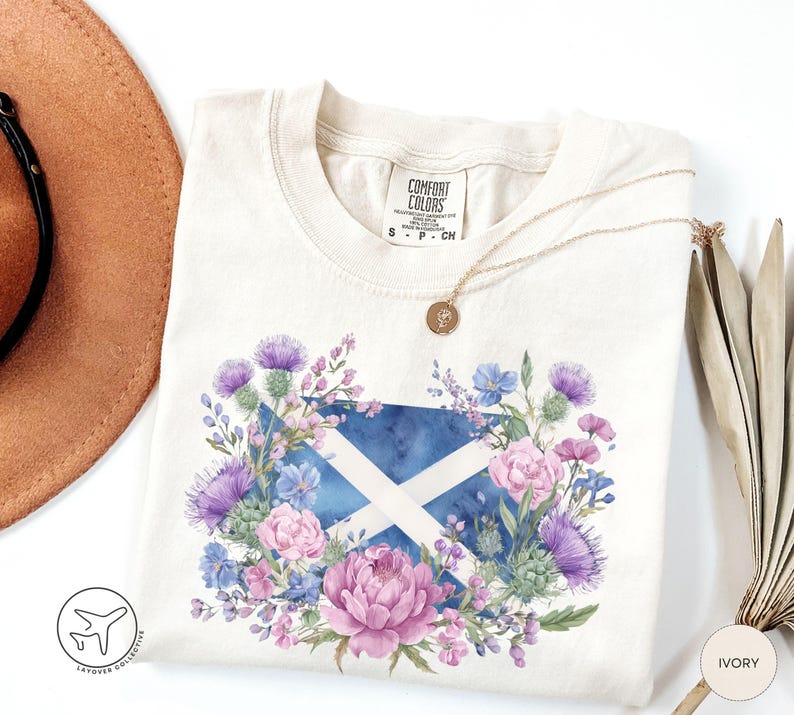 May include: Ivory-colored t-shirt featuring a watercolor design of the Scottish flag, surrounded by purple thistles, pink peonies, and blue flowers. A gold necklace and a brown hat are visible. The shirt has a Comfort Colors label.