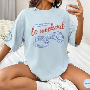 May include: Light blue t-shirt with the words "le weekend" in red script, along with the days of the week in French. A blue illustration of a coffee cup and croissant is also featured. The shirt is a casual, oversized style.