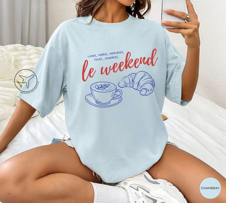 May include: Light blue t-shirt with the words "le weekend" in red script, along with the days of the week in French. A blue illustration of a coffee cup and croissant is also featured. The shirt is a casual, oversized style.