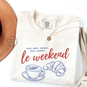 May include: Ivory-colored t-shirt with the words "le weekend" in red script, and the days of the week in French. A coffee cup and croissant are illustrated in blue. A gold necklace and a hat are also in the image.