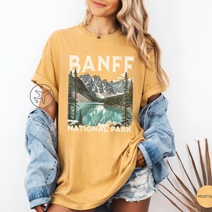 Może przedstawiać: Musztardowa koszulka z malowniczą ilustracją Parku Narodowego Banff. Grafika przedstawia jezioro, g&oacute;ry i drzewa, z napisem "BANFF NATIONAL PARK" białymi literami. Koszulka ma kr&oacute;tkie rękawy i luźny kr&oacute;j.