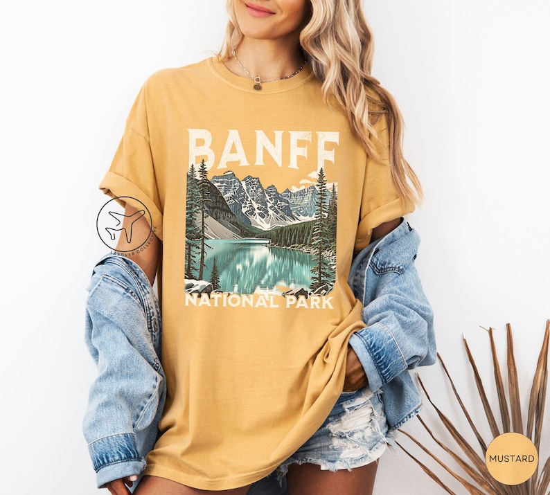 Może przedstawiać: Musztardowa koszulka z malowniczą ilustracją Parku Narodowego Banff. Grafika przedstawia jezioro, g&oacute;ry i drzewa, z napisem "BANFF NATIONAL PARK" białymi literami. Koszulka ma kr&oacute;tkie rękawy i luźny kr&oacute;j.
