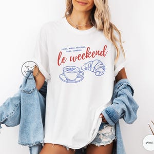 May include: White t-shirt with a graphic design featuring the text "le weekend" in red script, along with a blue illustration of a coffee cup and croissant. The shirt has short sleeves.