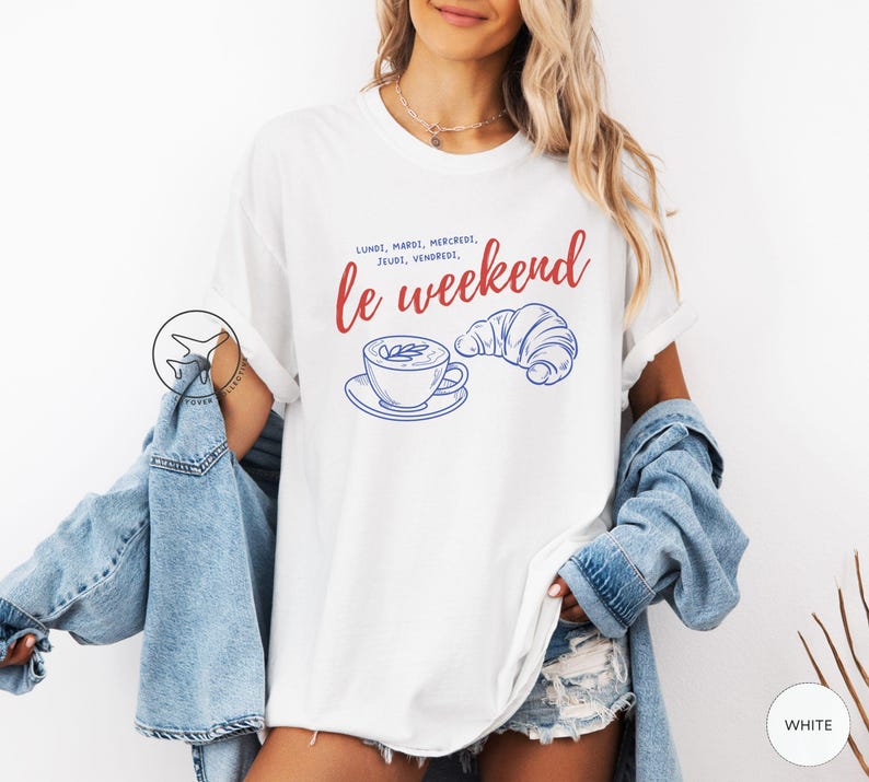 May include: White t-shirt with a graphic design featuring the text "le weekend" in red script, along with a blue illustration of a coffee cup and croissant. The shirt has short sleeves.