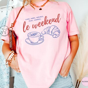 May include: A light pink t-shirt with the words "le weekend" in red script, along with the days of the week in French. A blue line drawing of a coffee cup and croissant are also featured on the shirt.