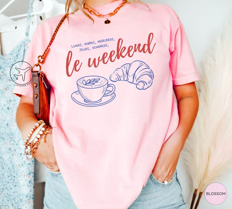 May include: A light pink t-shirt with the words "le weekend" in red script, along with the days of the week in French. A blue line drawing of a coffee cup and croissant are also featured on the shirt.