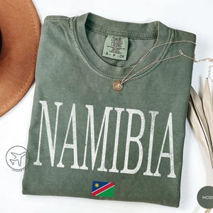 Peut inclure: T-shirt vert mousse avec le mot "NAMIBIA" en lettres blanches. Le t-shirt présente le drapeau namibien et une étiquette Comfort Colors. Un collier en or avec un pendentif repose sur le t-shirt.