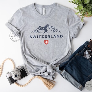 K&ouml;nnte beinhalten: Hellgraues T-Shirt mit einem marineblauen Bergmotiv und dem Wort "SCHWEIZ". Darunter ein rot-wei&szlig;es Schweizer Kreuz. Das T-Shirt wird mit einer blauen Jeans und einer Sonnenbrille kombiniert. Das T-Shirt ist aus Athletic Heather-Material.