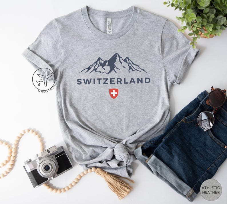 K&ouml;nnte beinhalten: Hellgraues T-Shirt mit einem marineblauen Bergmotiv und dem Wort "SCHWEIZ". Darunter ein rot-wei&szlig;es Schweizer Kreuz. Das T-Shirt wird mit einer blauen Jeans und einer Sonnenbrille kombiniert. Das T-Shirt ist aus Athletic Heather-Material.