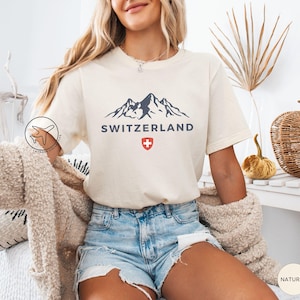 K&ouml;nnte beinhalten: Cremefarbenes T-Shirt mit einem dunkelblauen Bergmotiv, dem Wort "SWITZERLAND" und einem rot-wei&szlig;en Schweizer Kreuz. Das Shirt wird mit Jeansshorts kombiniert. Das Thema ist Reisen und Tourismus.