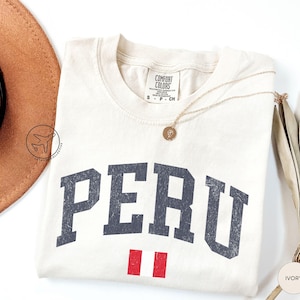 May include: Ivory-colored t-shirt with the word "PERU" in dark gray, arched lettering. Below the text is a small Peruvian flag. A gold necklace and a brown hat are also visible.