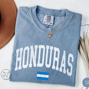 May include: Blue jean colored t-shirt with the word "HONDURAS" in white block letters. The Honduras flag is printed below the word. A gold necklace with a pendant is on the shirt.