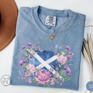 May include: Blue jean Comfort Colors t-shirt featuring a floral design around a Scottish flag. The design includes purple thistles, pink peonies, and blue flowers. A gold necklace with a pendant rests on the shirt.