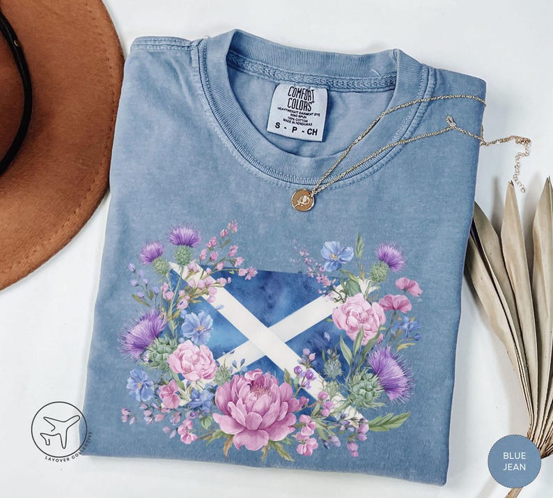 May include: Blue jean Comfort Colors t-shirt featuring a floral design around a Scottish flag. The design includes purple thistles, pink peonies, and blue flowers. A gold necklace with a pendant rests on the shirt.