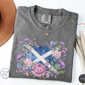 May include: A gray Comfort Colors t-shirt featuring a floral design with the Scottish flag at the center. The design includes a blue and white flag, surrounded by pink, purple, and blue flowers. A silver necklace with a small pendant rests on the shirt.