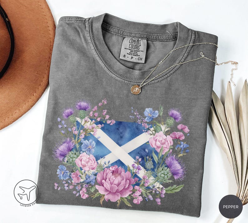 May include: A gray Comfort Colors t-shirt featuring a floral design with the Scottish flag at the center. The design includes a blue and white flag, surrounded by pink, purple, and blue flowers. A silver necklace with a small pendant rests on the shirt.