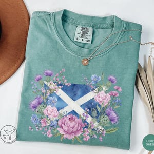 May include: Light green t-shirt featuring a floral design around a Scottish flag. The design includes purple thistles, pink peonies, and blue flowers. The shirt has a crew neck and short sleeves. A silver necklace with a pendant is draped on the shirt.