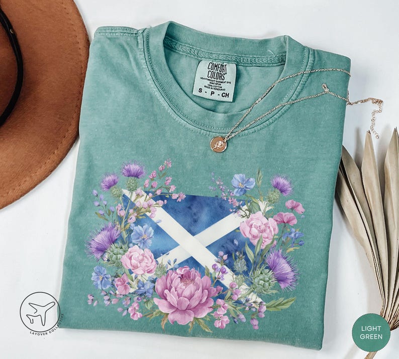 May include: Light green t-shirt featuring a floral design around a Scottish flag. The design includes purple thistles, pink peonies, and blue flowers. The shirt has a crew neck and short sleeves. A silver necklace with a pendant is draped on the shirt.