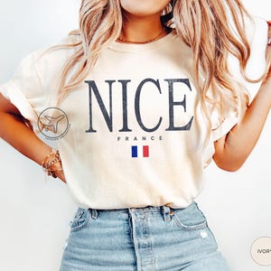 May include: Ivory-colored t-shirt with the word "NICE" in dark blue, distressed font. Below, "FRANCE" is printed, with a small French flag. The shirt is a casual style, suitable for travel or everyday wear.