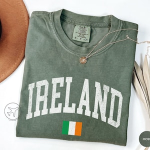 May include: Moss green t-shirt with the word "IRELAND" in white block letters and the Irish flag. The shirt has a crew neck and short sleeves. A gold necklace with a pendant is draped on the shirt.