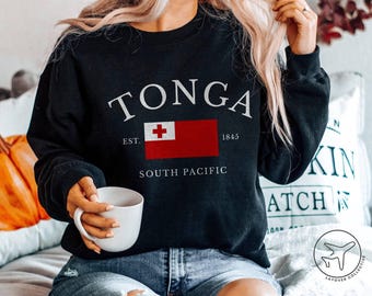 May include: Black sweatshirt featuring the word "TONGA" in white arched text. Below is a red rectangle with a white cross, the flag of Tonga. The text "EST. 1845" and "SOUTH PACIFIC" are also printed on the sweatshirt.