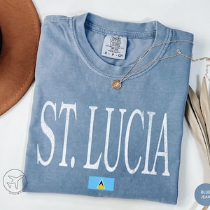 May include: A blue jean colored t-shirt with the text "ST. LUCIA" in white block letters. The shirt features a small St. Lucia flag graphic and a gold necklace. The shirt is made of a soft material.