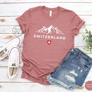 K&ouml;nnte beinhalten: Mauvefarbenes T-Shirt mit dem Wort "SCHWEIZ" und einer wei&szlig;en Berg-Grafik. Die Schweizer Flagge befindet sich unter dem Text. Wei&szlig;e Turnschuhe und Jeansshorts liegen neben dem Shirt.