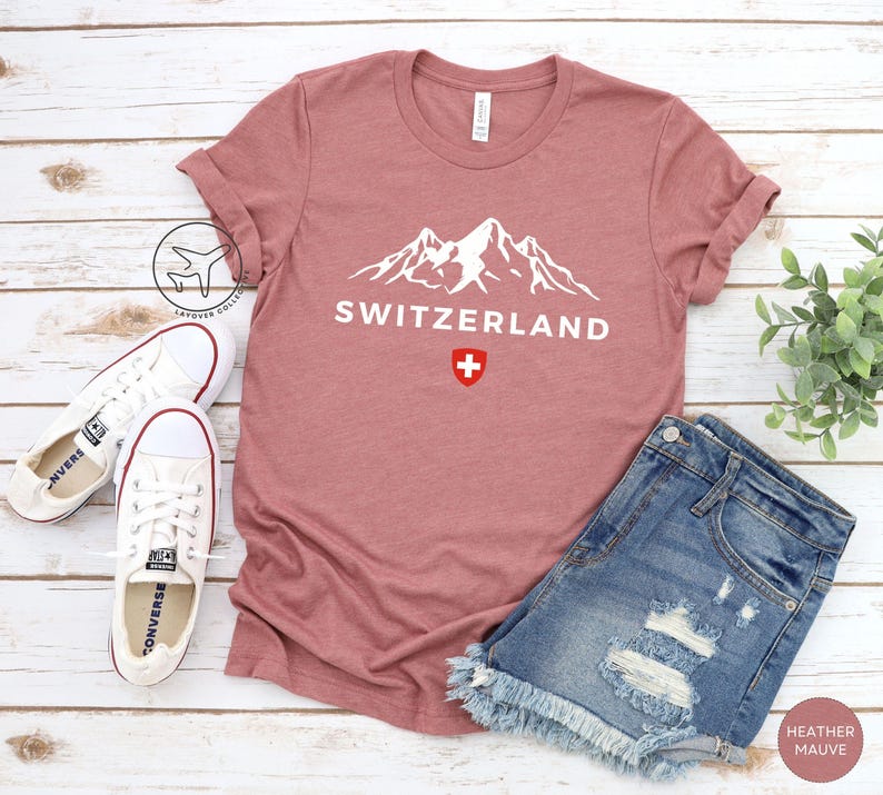 K&ouml;nnte beinhalten: Mauvefarbenes T-Shirt mit dem Wort "SCHWEIZ" und einer wei&szlig;en Berg-Grafik. Die Schweizer Flagge befindet sich unter dem Text. Wei&szlig;e Turnschuhe und Jeansshorts liegen neben dem Shirt.