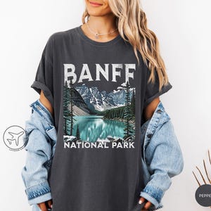 Może przedstawiać: Ciemnoszara koszulka z grafiką Parku Narodowego Banff. Projekt zawiera napis "BANFF" białymi, blokowymi literami nad ilustracją jeziora, g&oacute;r i drzew. Poniżej znajduje się napis "NATIONAL PARK".