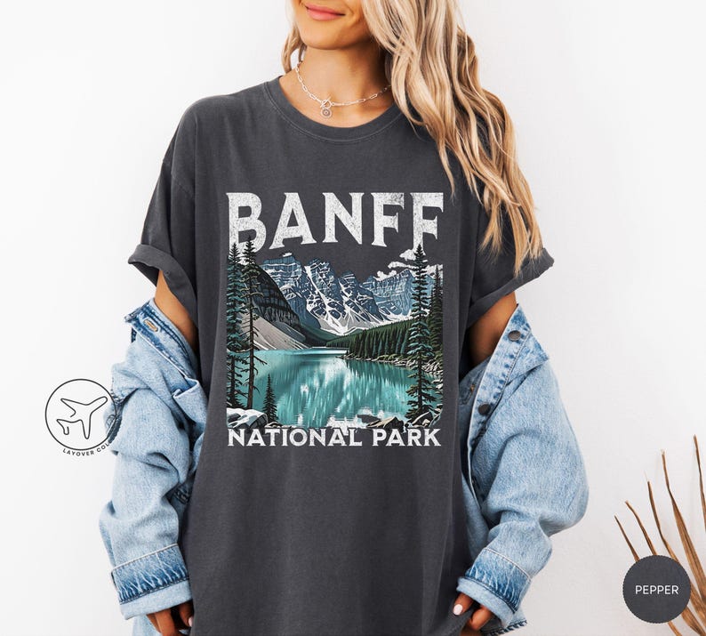 Może przedstawiać: Ciemnoszara koszulka z grafiką Parku Narodowego Banff. Projekt zawiera napis "BANFF" białymi, blokowymi literami nad ilustracją jeziora, g&oacute;r i drzew. Poniżej znajduje się napis "NATIONAL PARK".