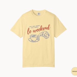 May include: Banana-colored t-shirt with the phrase "le weekend" in red script, accompanied by a blue illustration of a coffee cup and croissant. The shirt also features the days of the week in French.