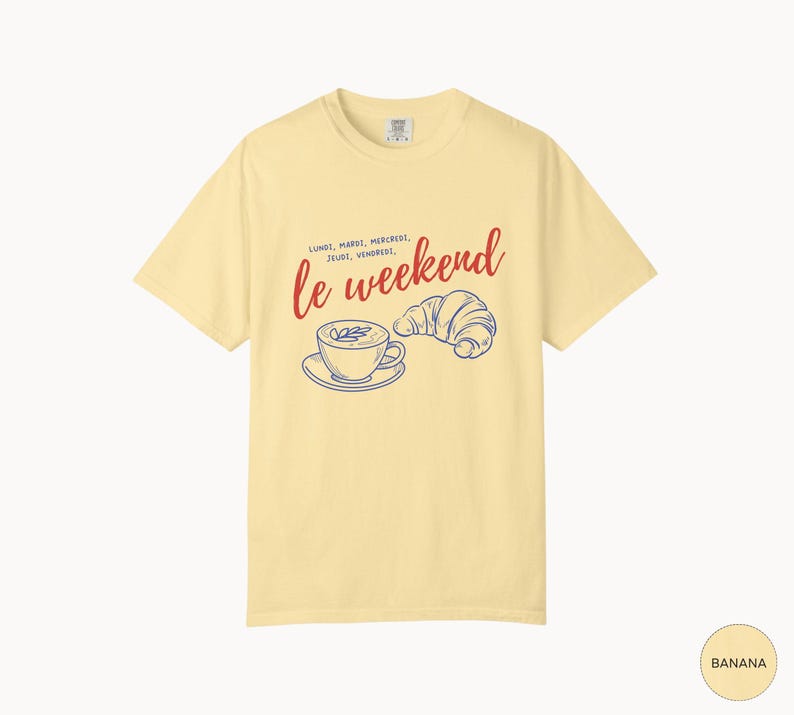 May include: Banana-colored t-shirt with the phrase "le weekend" in red script, accompanied by a blue illustration of a coffee cup and croissant. The shirt also features the days of the week in French.
