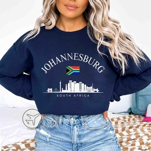 May include: Navy blue sweatshirt featuring the city name "JOHANNESBURG" in white arched text above a skyline graphic. The South African flag is displayed next to the city name. The text "SOUTH AFRICA" is below the skyline.