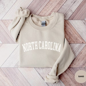 May include: A sand-colored sweatshirt with the text "NORTH CAROLINA" in white, arched lettering. The sweatshirt is folded and laid on a woodgrain background.