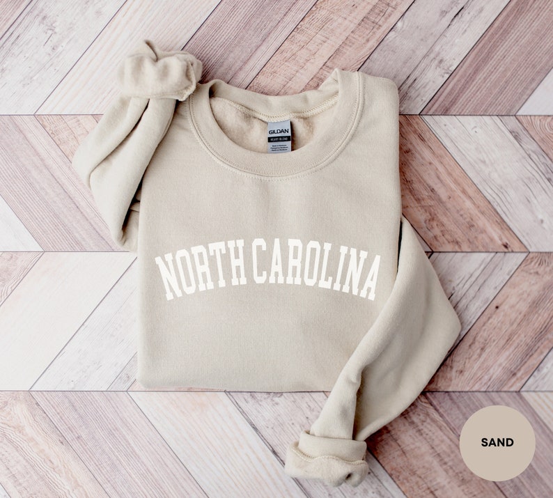 May include: A sand-colored sweatshirt with the text "NORTH CAROLINA" in white, arched lettering. The sweatshirt is folded and laid on a woodgrain background.
