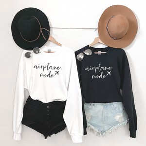 May include: Two white and black sweatshirts with the text "airplane mode" and a small airplane graphic. The sweatshirts are hanging on hangers and are styled with hats and shorts.