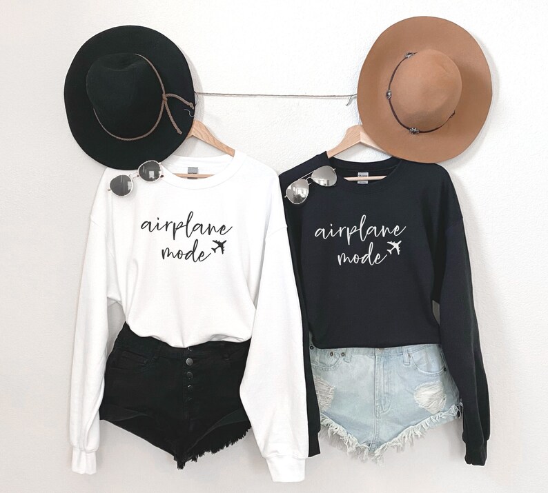 May include: Two white and black sweatshirts with the text "airplane mode" and a small airplane graphic. The sweatshirts are hanging on hangers and are styled with hats and shorts.
