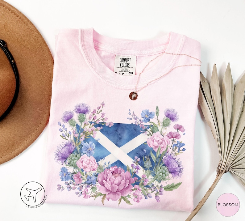 May include: A light pink Comfort Colors t-shirt featuring a floral design with the Scottish flag at the center. The design includes purple thistles, pink peonies, and blue flowers. A gold necklace with a small pendant is visible.