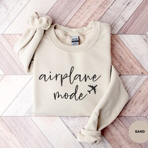 May include: A beige sweatshirt with the text "airplane mode" and a small airplane icon printed on the front. The sweatshirt is folded and laid on a wooden surface.