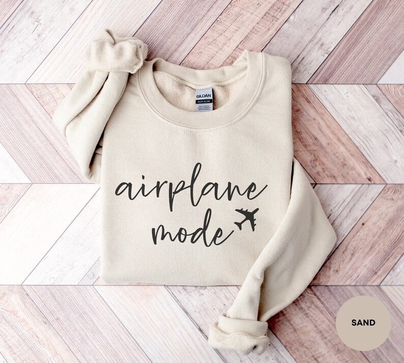 May include: A beige sweatshirt with the text "airplane mode" and a small airplane icon printed on the front. The sweatshirt is folded and laid on a wooden surface.