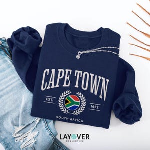 Könnte beinhalten: Ein marineblaues Sweatshirt mit dem weißen Schriftzug "CAPE TOWN", der südafrikanischen Flagge und dem Text "SOUTH AFRICA". Das Sweatshirt zeigt auch "EST. 1652". Eine silberne Halskette ist über dem Sweatshirt drapiert.
