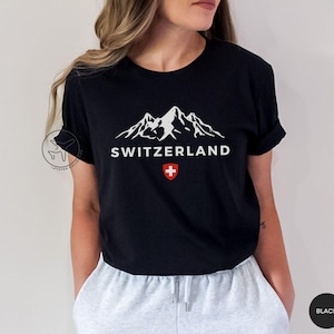 K&ouml;nnte beinhalten: Schwarzes T-Shirt mit wei&szlig;er Berglandschaft und dem Wort "SWITZERLAND". Darunter ein rot-wei&szlig;es Schweizer Kreuz. Das T-Shirt hat einen Rundhalsausschnitt.