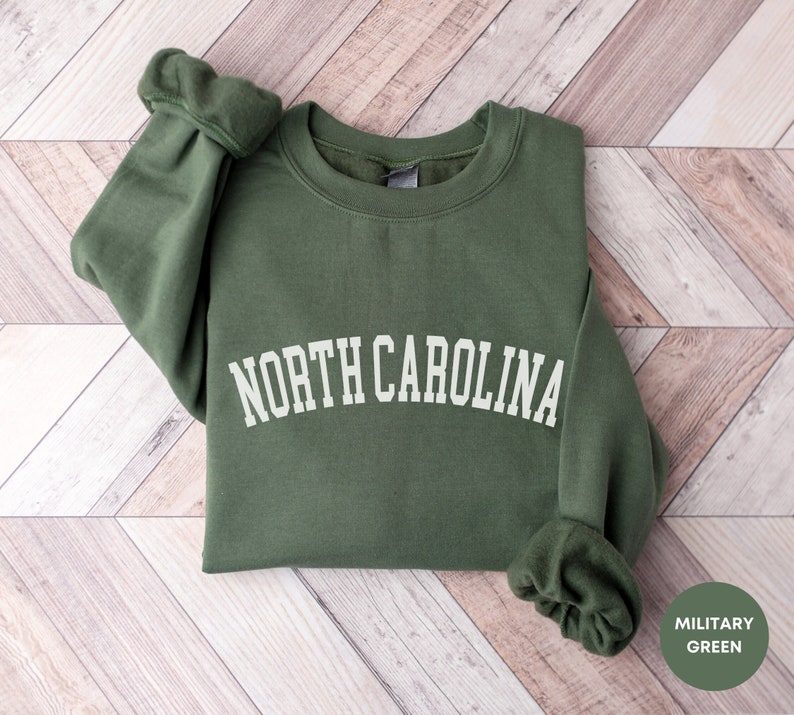 May include: A green sweatshirt with the text "NORTH CAROLINA" in white lettering. The sweatshirt is folded and laid on a woodgrain surface. The text "MILITARY GREEN" is visible in the lower right corner of the image.