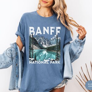 Może przedstawiać: Niebieska koszulka z napisem "BANFF" w białych literach blokowych nad malowniczą ilustracją jeziora i g&oacute;r. Poniżej ilustracji znajdują się słowa "NATIONAL PARK". Koszulka jest w jednolitym niebieskim kolorze.