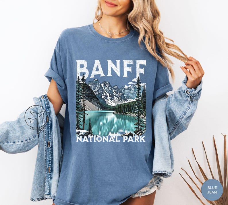 Może przedstawiać: Niebieska koszulka z napisem "BANFF" w białych literach blokowych nad malowniczą ilustracją jeziora i g&oacute;r. Poniżej ilustracji znajdują się słowa "NATIONAL PARK". Koszulka jest w jednolitym niebieskim kolorze.
